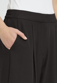 Pantalones negros plisados con textura suave, con cintura elástica con detalle fruncido y bolsillos laterales.