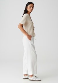 Beige short-sleeve top with textured fabric and tie detail, paired with wide white pants that are cuffed at the hem. White sneakers complete the look.