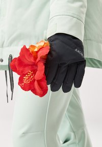 Svarta vinterhandskar med en texturerad yta, hållna i en hand bredvid en livfull röd och orange hibiscusblomma, mot en mintgrön jacka.