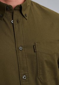 Camicetta verde oliva con bottoni realizzata in tessuto testurizzato. Presenta una tasca sul petto, bottoni di colori contrastanti e un'etichetta del marchio sulla tasca.