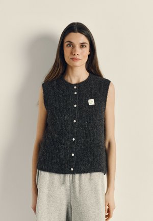Gilet sans manches gris foncé en tissu texturé, avec un devant boutonné et une petite étiquette sur le côté gauche.