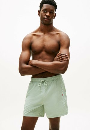 Mężczyzna bez koszulki z założonymi skrzyżowanymi ramionami, ubrany w jasnooliwkowe spodenki kąpielowe Tommy Hilfiger na jednolitym białym tle.