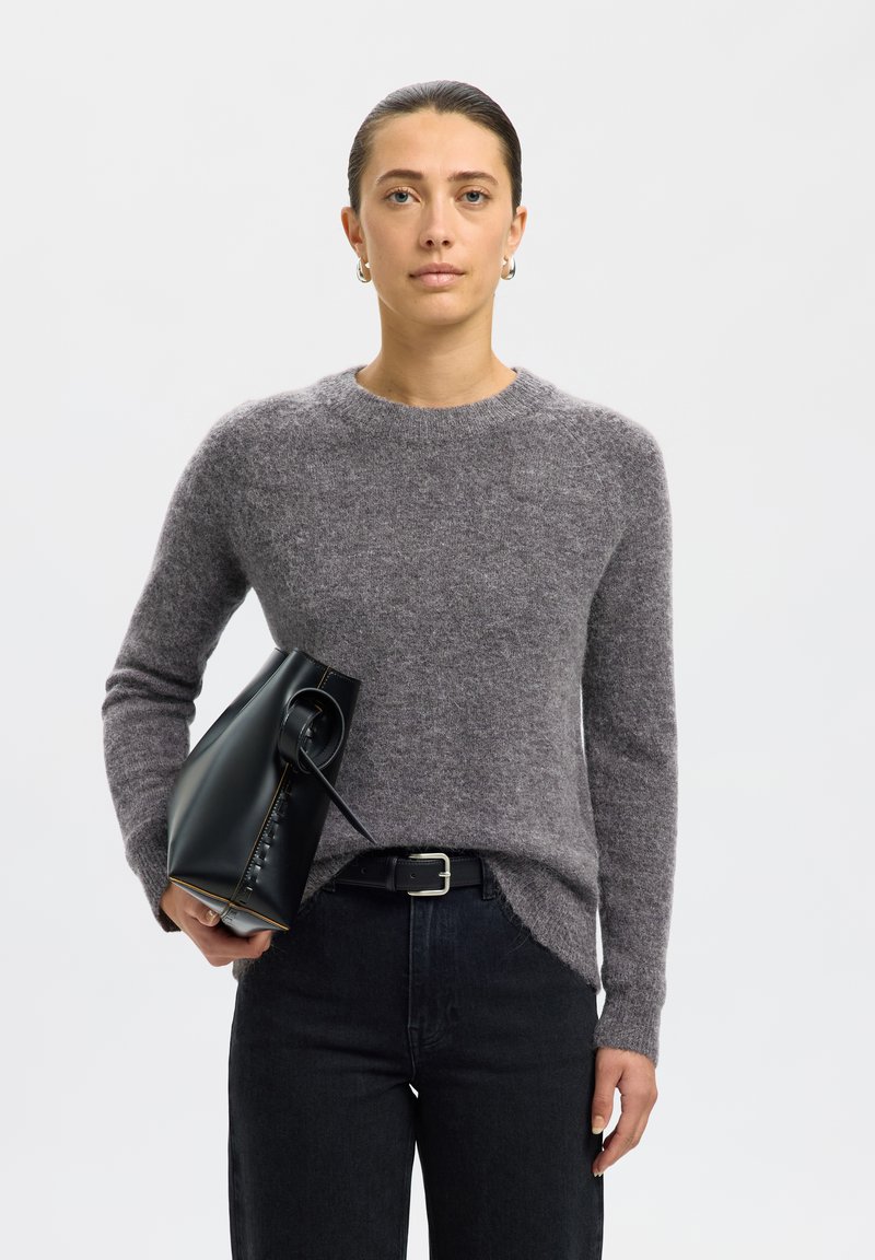 Kvinde med glat tilbagefejet hår iført en grå sweater og sorte jeans, der holder en sort læderhåndtaske mod en ensfarvet baggrund.