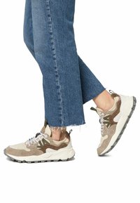 Beige und braune Sneaker aus Wildleder und Mesh-Materialien. Sie verfügen über eine strukturierte Sohle und einen Schnürverschluss, der durch grüne Akzente hervorgehoben wird.