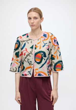 Femme aux cheveux blonds attachés portant une veste crème avec une broderie florale colorée et un pantalon bordeaux taille haute, debout devant un fond uni.
