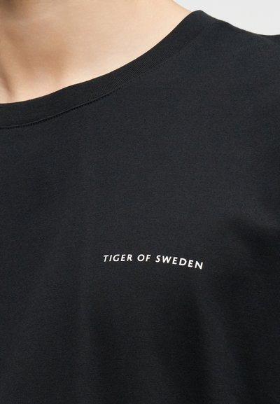 Κοντινό πλάνο ενός ατόμου που φοράει μαύρο πουκάμισο με στρογγυλή λαιμόκοψη και λευκό κείμενο "Tiger of Sweden" στο στήθος.