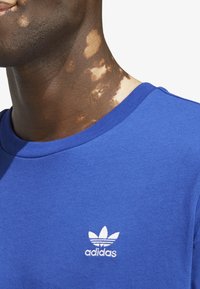 Camiseta de algodón azul con cuello redondo, mangas cortas y un logo de adidas bordado en blanco en el lado izquierdo del pecho.