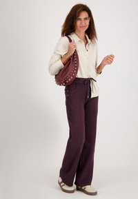 Vrouw in een crèmekleurig blouse met knopen, donkere paarse broek, beige sneakers en met een gestikte burgundy schoudertas, staand voor een witte achtergrond.