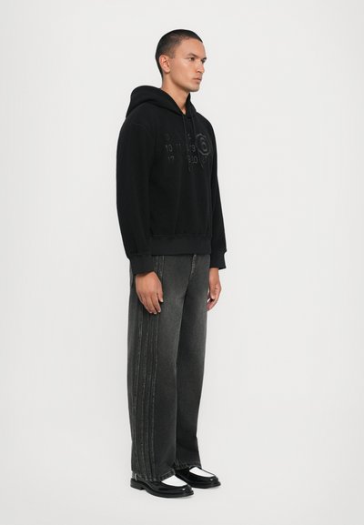 Svart huvtröja med präglade grafiska detaljer, kombinerad med löst sittande mörkgrå jeans med vertikala sömmar. Svart och vita skor.