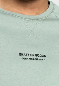 Sudadera verde desvaída hecha de algodón suave con cuello redondo acanalado; presenta texto bordado en negro: "PRODUCTOS ARTESANALES - PIER ONE DENIM."