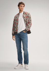 Jeune homme debout avec une main dans la poche, portant une veste bomber à motifs, un t-shirt blanc, un jean bleu et des baskets blanches.