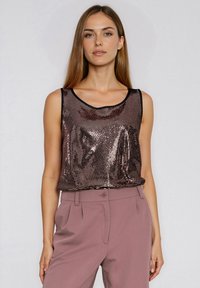 Kobieta o prostych, jasnobrązowych włosach, ubrana w bezrękawnik z metalicznych cekinów i wysokowalne mauve spodnie, stojąca na tle jednolitej ściany.