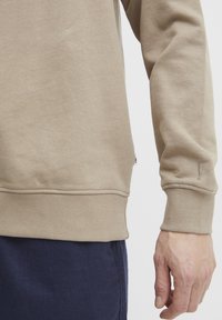 Beige sweatshirt made of soft cotton, featuring a ribbed hem and cuffs. Hand in dark pants rests beside. Minimal stitching and no visible logos.