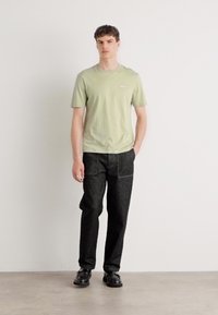 Lys grøn kortærmet t-shirt, parret med mørke, strukturerede bukser og sorte sko. Modellen står foran en ensfarvet hvid baggrund.