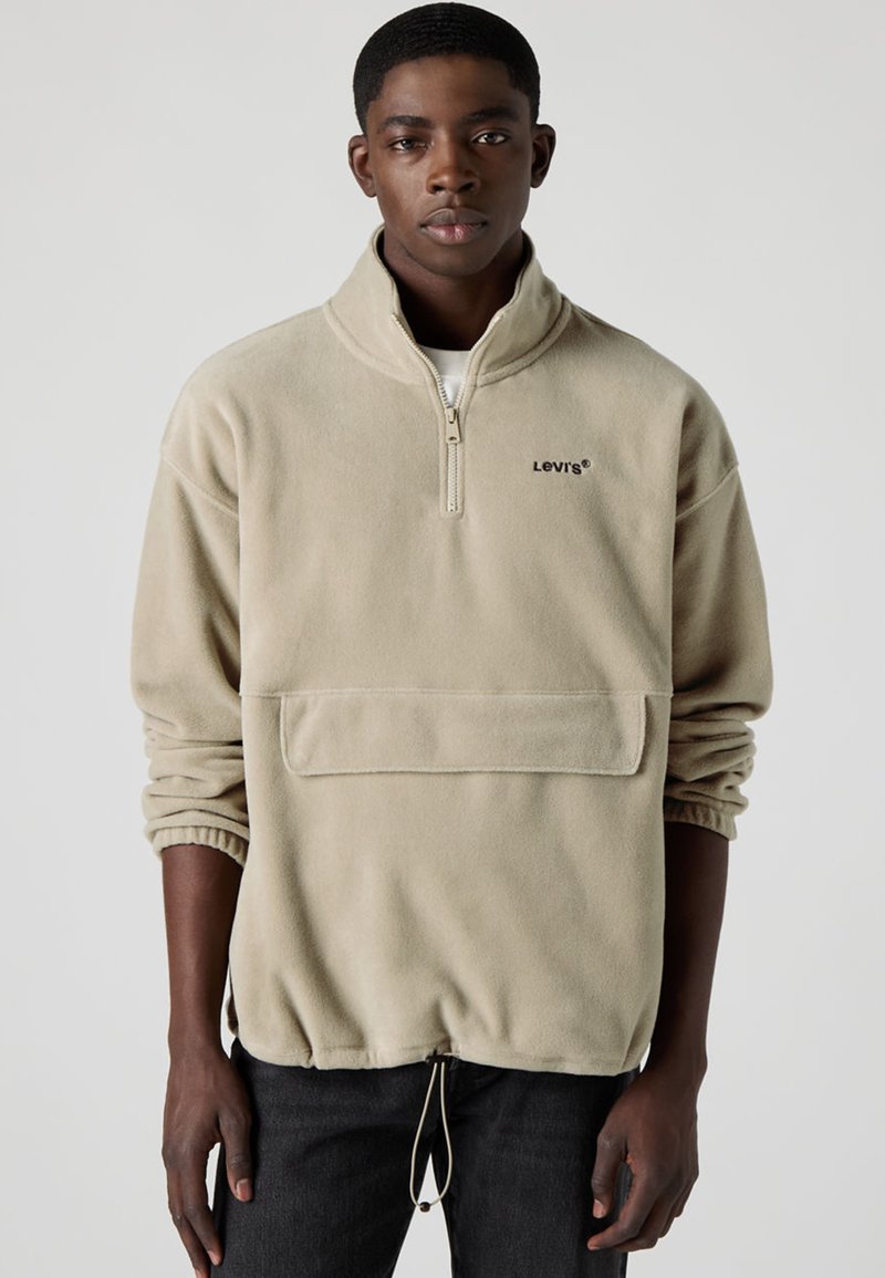 Beige fleecetröja med halvblixtlås, elastiska muddar och framficka. Har dragsko i nederkant och broderad logotyp på bröstet.