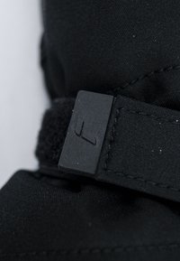 Cinghia in tessuto nera con chiusura Velcro e una toppa di gomma rettangolare con logo in rilievo. Il materiale ha una superficie liscia.