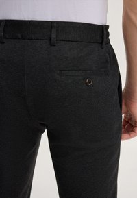 Pantaloni neri in tessuto testurizzato, con tasca sul retro dotata di bottone e piccolo etichetta del marchio sulla cintura.