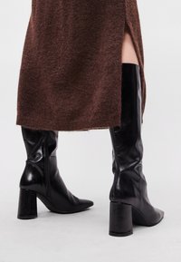 Melnas ādas līdz-ceļgala kurpes ar šķautņu purngaliem un blokveida papēdi, ar gludu tekstūru un smalku rāvējslēdzēju detaļu sānos.