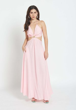 Ženska z dolgimi temnimi lasmi nosi sleeveless roza maxi obleko s halter ovratnikom, izrezi in zlato zaponko, stoji z eno roko v žepu.