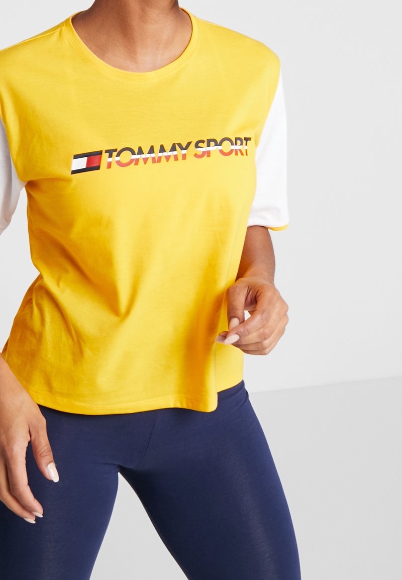 Žluté bavlněné tričko s krátkým rukávem a bílými rukávy, s logem "TOMMY SPORT" v červené a černé barvě. Hladká textura, volný střih.