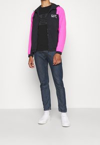 Färgglad vindjacka med svart kropp och rosa ärmar, med huva och dragkedja. I kombination med en svart t-shirt och blå jeans.