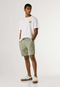 Moški stoji z rokami v žepih, oblečen v belo majico, svetlo zelene kratke hlače in bele superge na enobarvnem ozadju.