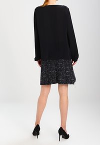 Schwarze Bluse mit langen Ärmeln über einem strukturierten schwarz-weißen gewebten Rock, kombiniert mit schwarzen High-Heels.
