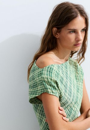 Fiatal nő zöld kockás rövid ujjú felsőben, vállával kibuggyanva, keresztbe font karokkal, gondolkodóan oldalra néz egy egyszerű háttér előtt.