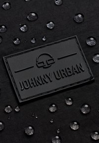Černá tkanina s texturovaným povrchem a kapkami vody. Vyvýšená, obdélníková černá etiketa s vyraženým nápisem "JOHNNY URBAN" uprostřed.
