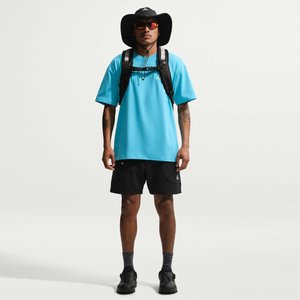 Man met een zwarte hoed, rode zonnebril, blauw T-shirt, zwarte korte broek, grijze sokken, zwarte schoenen en zwarte rugzak, staand tegen een witte achtergrond.