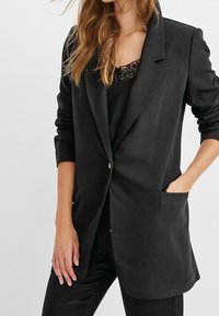 Sort blazer med en enkelt knap, to frontlommer og opsmøgede ærmer. Fremstillet af et glat, struktureret stof. Laget over en sort top.