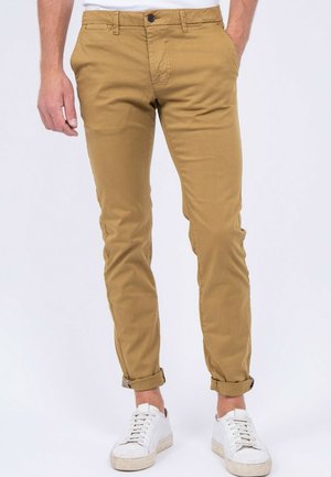 Homme portant un pantalon slim beige retroussé et des baskets blanches, debout devant un fond clair uni.