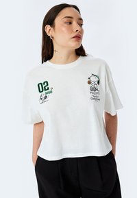 Camiseta de algodón blanca con mangas cortas, que presenta impresiones gráficas en verde y negro de Snoopy y detalles de texto en el frente. Diseño recortado.