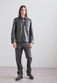 Black leather shirt jacket with dual chest pockets, paired with dark gray loose-fitting jeans and black ankle boots. Plain background.