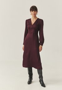 Rochie lungă burgundy cu decolteu adânc în V, mâneci bufante și talie ajustată. Asortată cu cizme negre, înguste, până la genunchi. Textură de țesătură netedă.