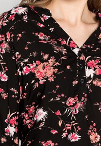 Blouse noire à fleurs en tissu doux, avec un col en V, une fermeture à boutons, et des motifs de fleurs roses et blanches. Détails incluant des volants subtils.