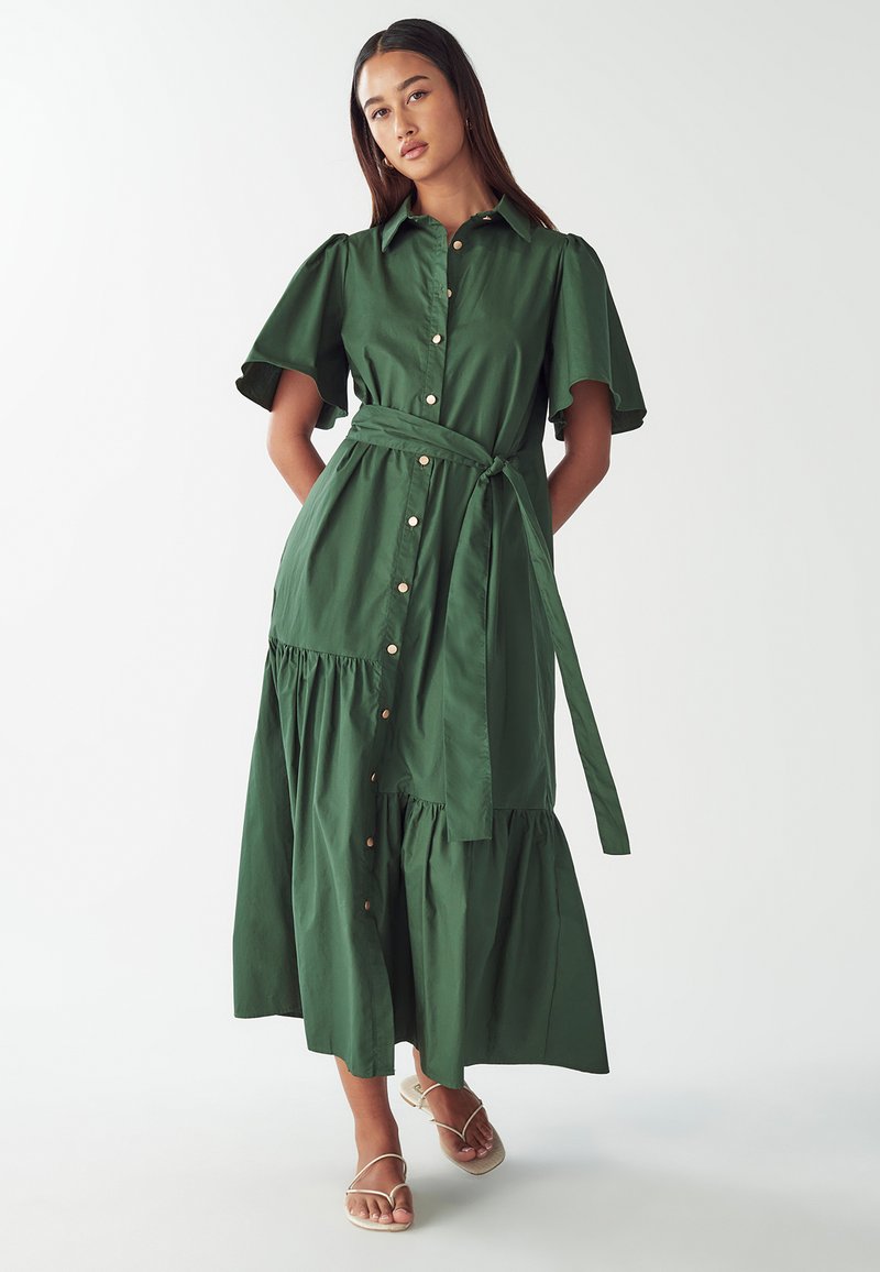 Vestito camicia in cotone verde con maniche corte a volant, davanti con bottoni e cintura in vita. Presenta una gonna a strati e bottoni dorati.