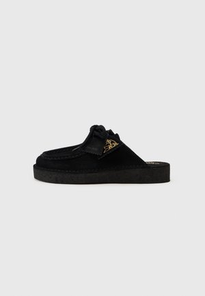 Chaussure glissante en daim noir avec une semelle moulée, comportant un détail en nœud et un accent logo doré sur le côté. Surface texturée avec des bords cousus.