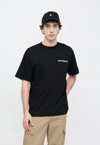 Czarny bawełniany T-shirt z krótkimi rękawami i okrągłym dekoltem, z białym logo Carhartt na lewej piersi. Noszony z beżowymi spodniami typu cargo.