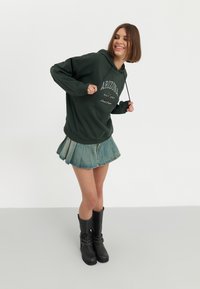 Donker groene hoodie met de tekst "ARIZONA" in het wit, gecombineerd met een denim plissérok en zwart kniehoge laarzen met zilveren accenten.