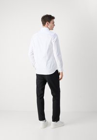 Camicia lunga bianca a maniche lunghe con polsini a bottoni, abbinata a pantaloni neri e sneakers bianche. Tessuto morbido, vestibilità su misura e colletto rigido.