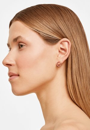 Roségoldene, runde Ohrringe mit glatter Oberfläche, bestehend aus zwei ineinandergreifenden Kreisen, getragen von einem Model mit langen braunen Haaren.