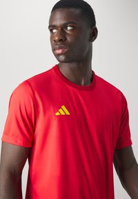 Rode sportieve shirt met korte mouwen, ronde hals, gemaakt van lichtgewicht materiaal; heeft een groen en geel Adidas-logo op de borst.
