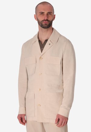 Uomo che indossa una giacca beige con bottoni e quattro grandi tasche frontali sopra una camicia marrone, in piedi davanti a uno sfondo semplice.