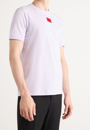 Man wearing light purple short-sleeve t-shirt with small red square logo on chest, paired with black pants against a plain background.
