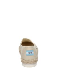 Beige canvas slip-on shoe with rope detailing around the sole and a light blue label on the heel reading "TOMS." Minimalist design.