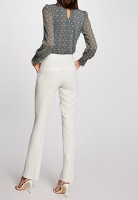 Luftige Bluse mit geometrischem Druck in dunklen Tönen, kombiniert mit cremefarbenen, maßgeschneiderten Hosen. Besitzt einen Schlitz im Rücken und elegante, hochhackige Sandalen.