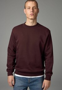 Sudadera burdeos con cuello redondo, puños y dobladillo acanalados, fabricada en tejido suave, llevada sobre una camiseta blanca, combinada con jeans de mezclilla.