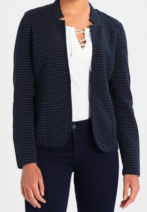 Kvinde iført en marineblå blazer med hvide polkaprikkker over en hvid snørelukningstop og mørke bukser, stående mod en hvid baggrund.