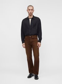 Homme portant une chemise noire à manches longues, un pantalon marron et des chaussures habillées noires, debout devant un fond clair uni.
